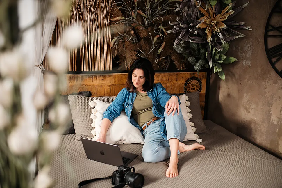 Rita laptop előtt ülve, ágyon dolgozik, mellette fényképezőgép.